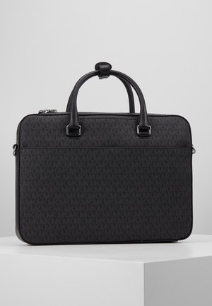 Mallette rectangulaire noire avec surface texturée et double poignée arrondie posée sur un piédestal blanc sur un fond gris clair.