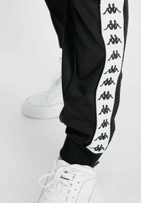 Pantalones atléticos negros fabricados con material suave, con una franja lateral blanca que presenta un logotipo negro repetido; combinados con zapatillas blancas.