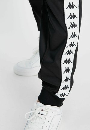 Pantaloni sportivi neri realizzati in materiale liscio, con una striscia laterale bianca e logo nero ripetuto; abbinati a sneakers bianche.