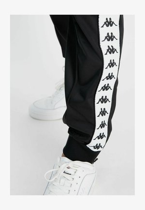 Pantaloni sportivi neri realizzati in materiale liscio, con una striscia laterale bianca e logo nero ripetuto; abbinati a sneakers bianche.