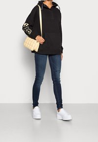 Zwarte hoodie met een voorzak en een capuchon, voorzien van grafische ontwerpen op de mouwen. Draagt donkerblauwe jeans en witte platform sneakers.