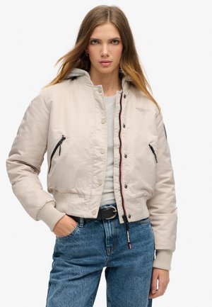 Veste bomber beige raccourcie avec une poche zippée, des poignets côtelés et une capuche grise. Présente des boutons pression et un détail de marque subtil.
