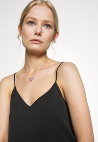 Czarny bezrękawnik z dekoltem w kształcie litery V, wykonany z gładkiej tkaniny. Uzupełniony srebrnym naszyjnikiem z zawieszką w kształcie serca.