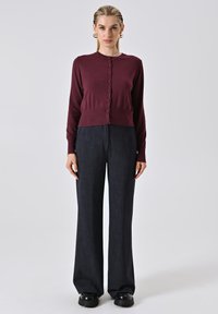 Frau steht und trägt eine geknöpfte burgunderfarbene Strickjacke, dunkle weite Hosen, schwarze klobige Schuhe und goldene Creolen vor einem weißen Hintergrund.