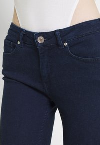 Jeans in denim blu scuro con design a vita media, tasche frontali, dettagli con bottoni argentati e tessuto liscio leggermente strutturato.