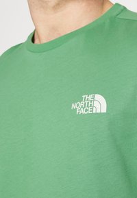 Green cotton t-shirt with a crew neck. Features a white logo print "THE NORTH FACE" on the left chest area. Smooth texture and casual fit.