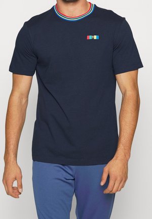 Mężczyzna ubrany w granatowy t-shirt z krótkim rękawem, wielokolorowym paskowanym kołnierzem i małą kolorową naszywką na klatce piersiowej.