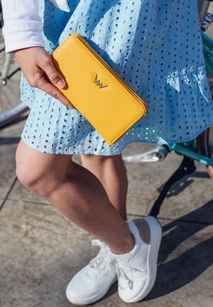 Yellow leather wallet with a zip closure, featuring a silver logo, held in a hand against a blue eyelet dress and a bicycle background.
