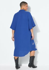 Robe bleue, longueur genou, avec une coupe décontractée, des manches courtes, un tissu texturé et des motifs de soleil brodés à l' ourlet. Portée avec des bottes de cow-boy noires.