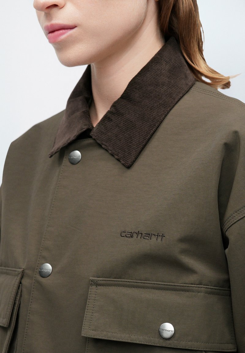 Olivgrüne Jacke mit braunem Cordkragen, ausgestattet mit silbernen Druckknöpfen und Taschenklappen; mit dem Markennamen 'Carhartt' auf der Brust gestickt.