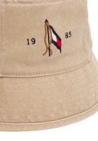 Beige bucket hat made of textured fabric, featuring embroidered logo with a flag design in red, white, and blue, and the year "1985."