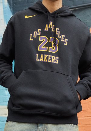 Sudadera negra con capucha con logo amarillo de Nike, texto "Los Angeles Lakers" y número 23 en púrpura y amarillo, llevada por una persona con las manos en los bolsillos.