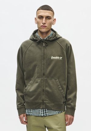 Jeune homme portant un sweat à capuche zippé vert olive, une chemise à carreaux en dessous et un pantalon kaki, debout devant un fond uni.