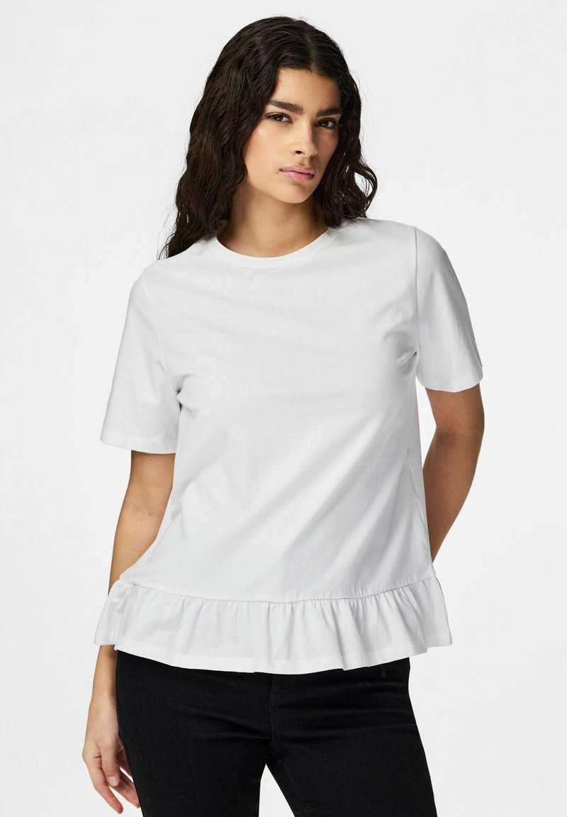 Femme aux cheveux longs, ondulés et bruns, portant un haut blanc uni à manches courtes avec un ourlet à volants et un pantalon noir, debout avec une main derrière le dos.