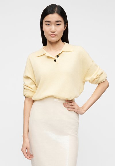 Svjetlo žuta, široka majica s ovratnikom i dva tamna gumba. Nošena preko sjajne, kremaste pencil suknje. Glatka tekstura.