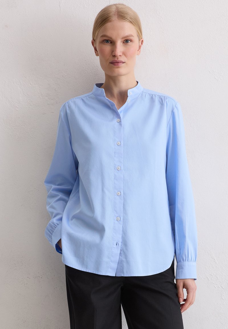 Blondynka w jasnoniebieskiej koszuli z mandaryńskim kołnierzykiem zapinanej na guziki oraz w czarnych spodniach, stojąca na tle gładkiej białej ściany.
