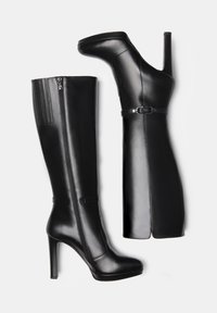 Bottes noires montantes en cuir verni, à bout pointu, avec un haut talon stiletto et une fermeture éclair sur le côté. Détails avec une fine sangle à la cheville.