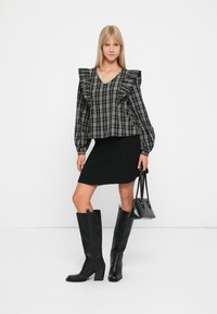 Blouse à carreaux noir et blanc avec épaules ruffles, associée à une jupe noire, des bottes hautes noires et un petit sac à main noir.