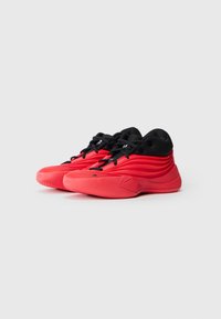Des chaussures de sport basses rouges et noires avec des lacets noirs et des semelles texturées, présentées sur un fond blanc uni, inclinées vers la gauche.