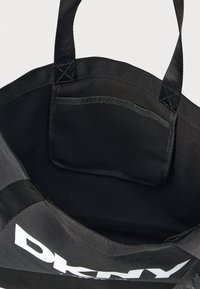 Saco de lona preto com alças duplas, bolso interior e logo DKNY branco proeminente de um lado. Textura suave, design simples.