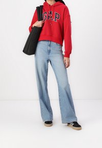 Czerwona bluza z kapturem z logo "GAP", luźny fason; jasnoniebieskie, wysokie, dzwonowe dżinsy; czarna torba; czarno-białe sneakersy z gumową podeszwą.