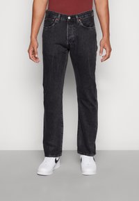 Jeans de mezclilla negros con corte recto, cinco bolsillos y costuras naranjas en contraste, combinados con zapatillas blancas.
