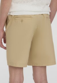 Pantaloni shorts beige realizzati in tessuto leggero con una texture liscia, dotati di una tasca posteriore con chiusura a bottone e una vestibilità rilassata.