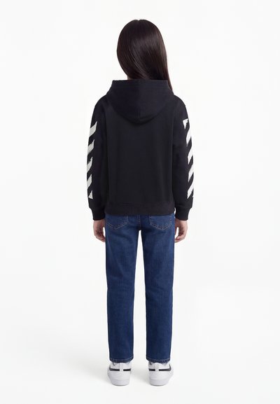 Kind met de rug naar de kijker, draagend een zwarte hoodie met witte diagonale strepen op de mouwen, een blauwe spijkerbroek en witte sneakers, op een witte achtergrond.