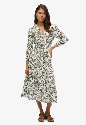 Vestido de manga larga en blanco con un patrón paisley floral verde, escote en V, botones en la parte delantera y detalle de falda en capas. Las sandalias completan el conjunto.