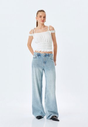 Top corto blanco con fruncido y tirantes caídos, combinado con vaqueros de pierna ancha azul claro. Unos zapatos planos negros de punta completan el atuendo.