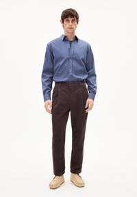 Outfit maschile che presenta una camicia di cotone blu e pantaloni di velluto a coste marrone scuro. Calzature slip-on beige completano il look.