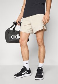 Shorts beiges et chaussures de sport noires avec des accents blancs. Un sac de sport noir avec le logo "adidas" blanc. Chaussettes blanches.