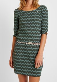 Zielona sukienka w zygzakowy wzór w niebieskim kolorze, z rękawami do trzech ćwiartek, okrągłym dekoltem i plecionym paskiem. Posiada guziki dekoracyjne na kieszeniach.