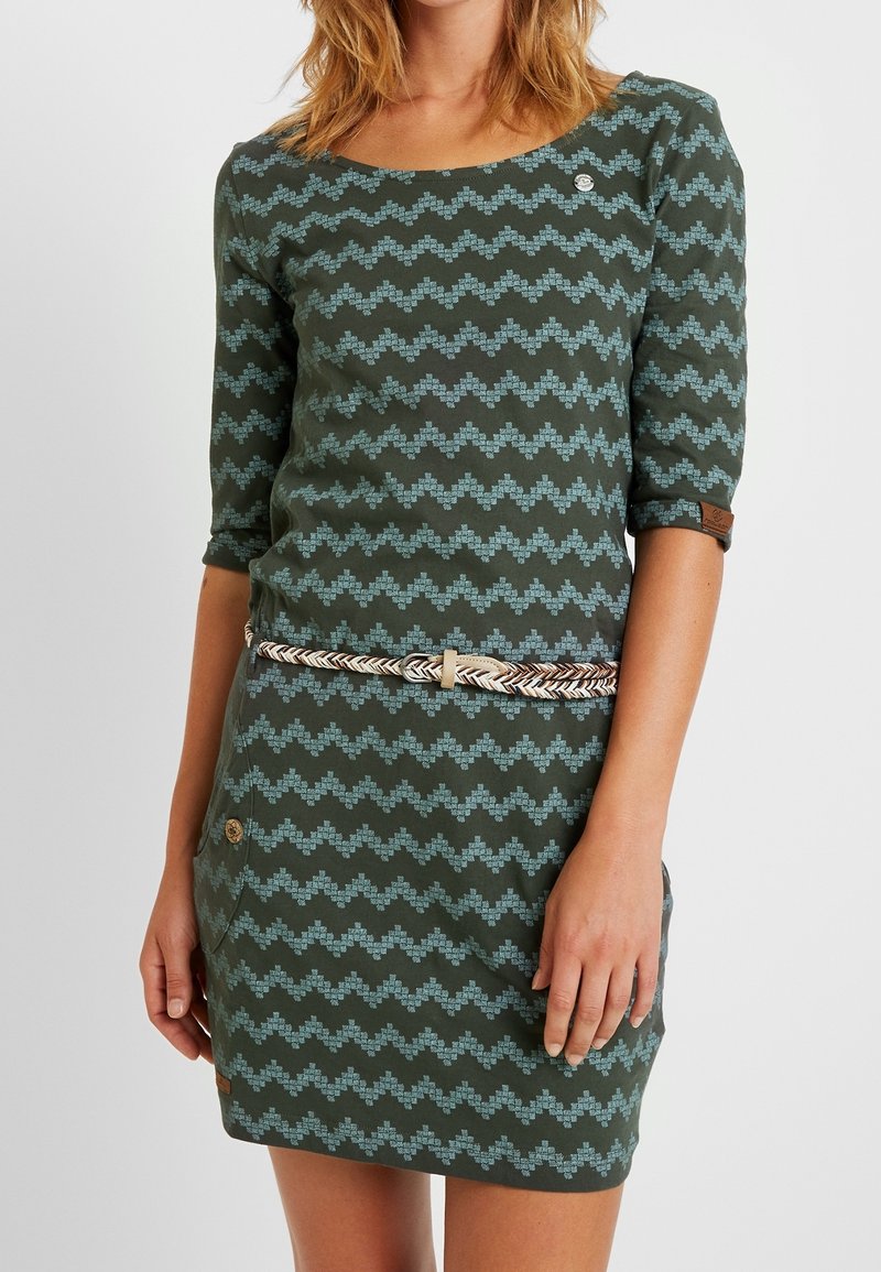 Zielona sukienka w zygzakowy wzór w niebieskim kolorze, z rękawami do trzech ćwiartek, okrągłym dekoltem i plecionym paskiem. Posiada guziki dekoracyjne na kieszeniach.