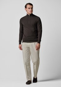 Suéter de punto marrón oscuro con cuello de cremallera hasta el pecho, combinado con pantalones beige claros y zapatos oscuros, modelo posando contra un fondo gris.