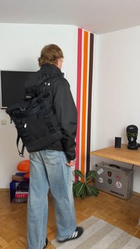 Svart ryggsekk med flere stropper og spenner, laget av slitesterkt materiale. Designet har et romslig hovedrom og teksturerte detaljer.