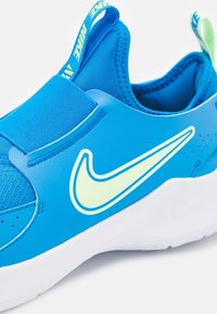 Blauwe sportieve schoen gemaakt van synthetisch materiaal met een getextureerd meshpaneel, voorzien van een groot geel Nike-logo, een elastische band en een trekhaak.