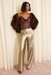 Donna che indossa un top in satin marrone, un'oversize giacca di pelliccia marrone scuro e pantaloni beige lucidi a vita alta e gamba larga, in piedi all'interno.