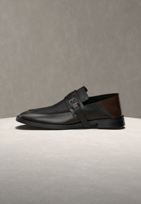 Mocassin en cuir noir avec détail marron au talon et une bride à boucle sur le dessus, présenté sur une surface claire sur un fond neutre.