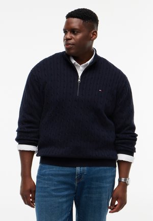 Homme portant un pull bleu marine à col zippé en maille torsadée par-dessus une chemise blanche aux manches retroussées et un jean bleu, regardant vers la gauche.