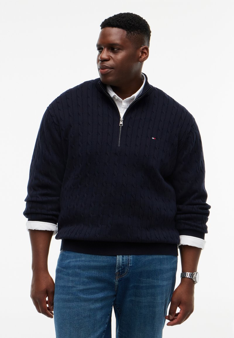 Homme portant un pull bleu marine à col zippé en maille torsadée par-dessus une chemise blanche aux manches retroussées et un jean bleu, regardant vers la gauche.
