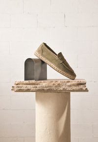 Mocassin espadrille en toile vert olive équilibré sur un bloc métallique courbé au sommet de dalles de pierre empilées sur un piédestal beige devant un mur en briques blanches.