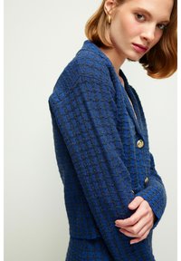 Blazer texturizado azul con un patrón de diamantes, diseño de doble botonadura y botones dorados. Ajuste entallado con hombros estructurados y mangas largas.