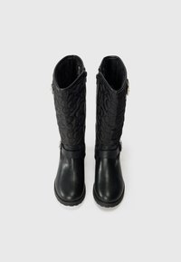 Botas negras hasta la rodilla con punta de cuero suave y caña de textil acolchado con un patrón decorativo, cierre lateral de cremallera y detalle de correa.