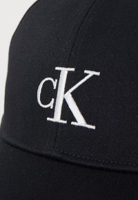 Casquette noire en mélange de coton avec une visière incurvée. Présente un logo "cK" brodé blanc à l'avant et une surface en tissu texturé.