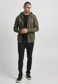 Olivengrønn hettegenser med glidelås og hvite snorer, kombinert med en svart skjorte og slim-fit svarte bukser. Teksturert stoff og sporty design.