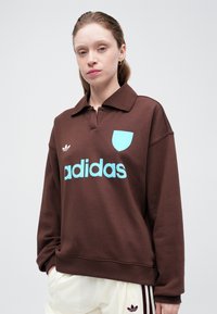 Rjav pulover s kapuco z ovratnikom, ki ima svetlo modro "adidas" logo in ujemajočo se emblemo na prsih. Mehka tekstura tkanine.
