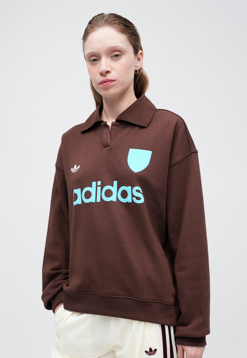 Sweatshirt pullover marron avec un col, arborant un logo "adidas" bleu clair et un emblème assorti sur la poitrine. Texture de tissu doux.