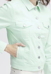 Chaqueta de mezclilla verde menta con botones plateados, dos bolsillos en el pecho y un diseño con cuello con detalles de costura a lo largo de las costuras.
