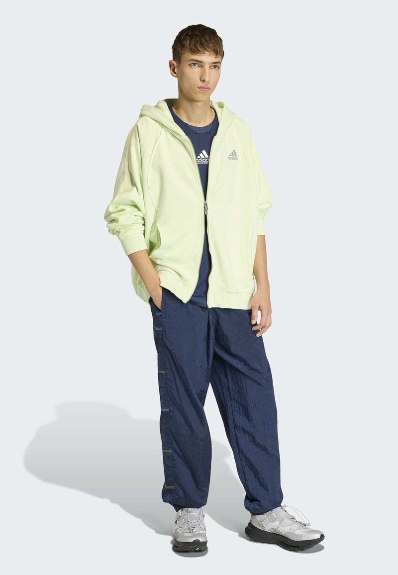 Sweat à capuche zippé vert clair, chemise marine, pantalon ample marine avec fermetures éclair latérales, et chaussures de sport grises, porté par une personne debout contre un fond clair.